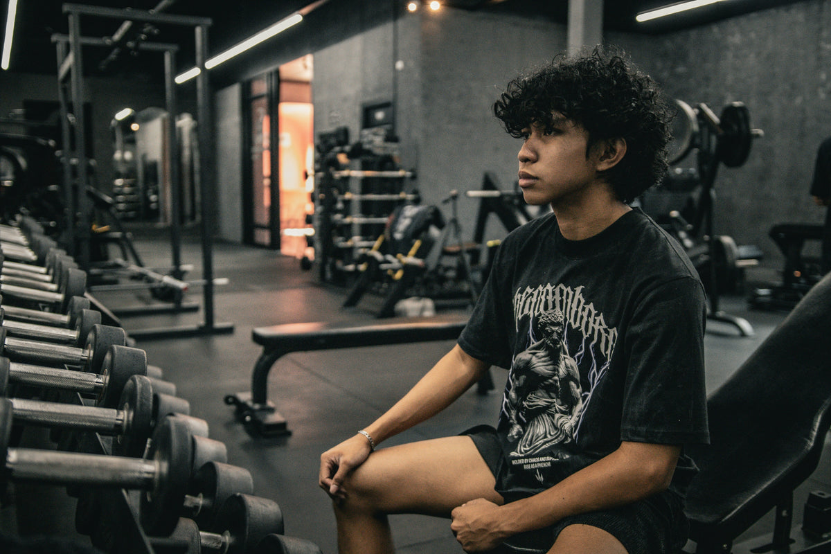 A young man sits on a gym bench, pausing before his workout as he considers whether creatine for beginners is worth trying before he's even started training.
