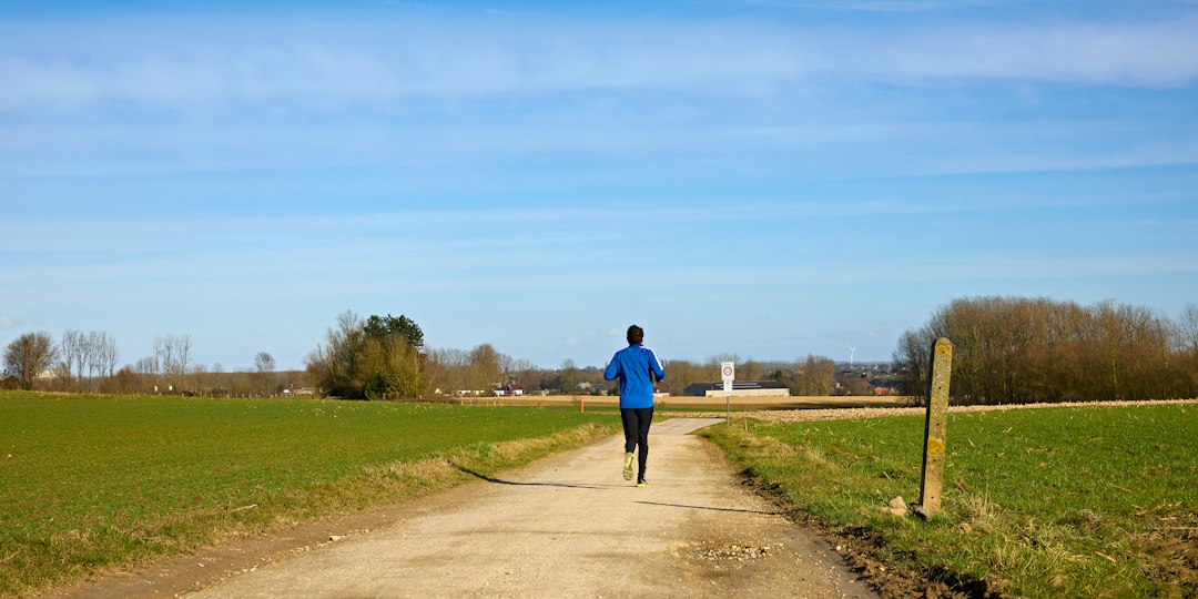 A male distance runner in athletic gear running along a dirt path through green fields, illustrating how runners need both hydration and electrolytes to sustain endurance training.