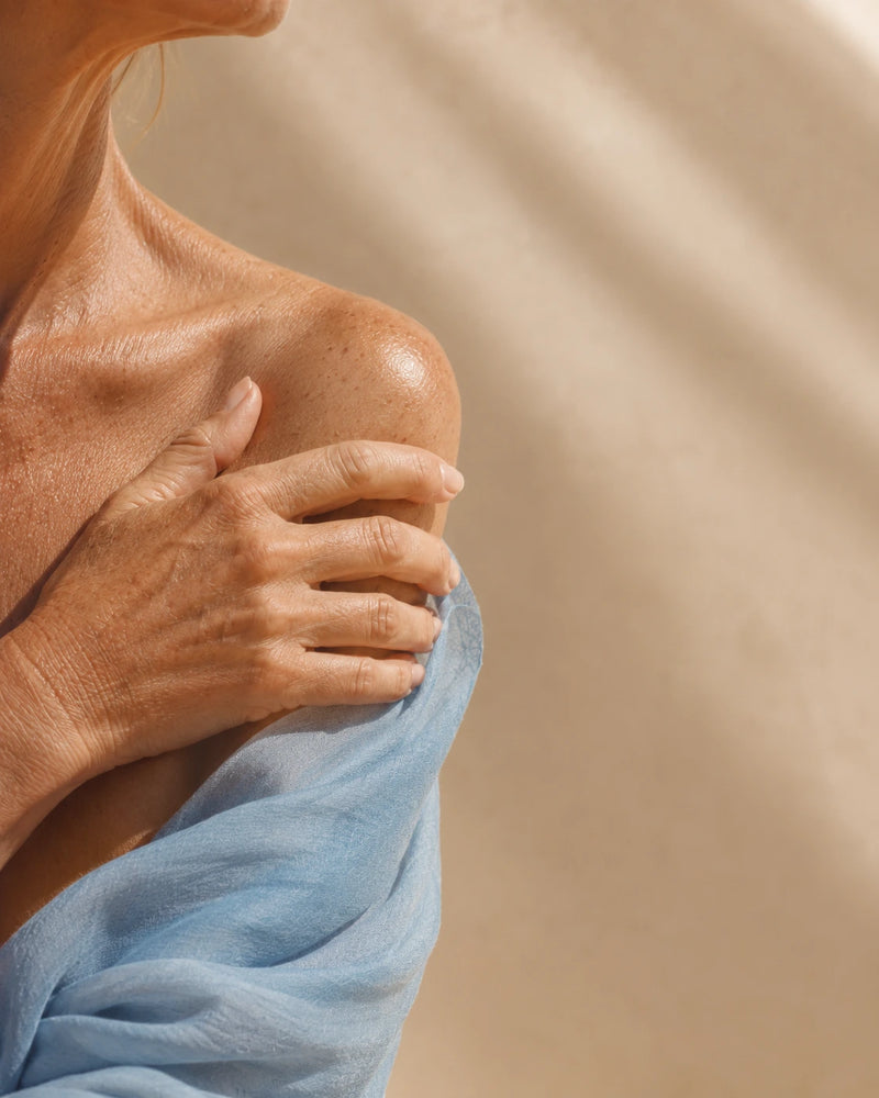 Close-up of a woman's shoulder and upper chest with her hand resting on her shoulder, wrapped in a light blue fabric against a warm beige background.