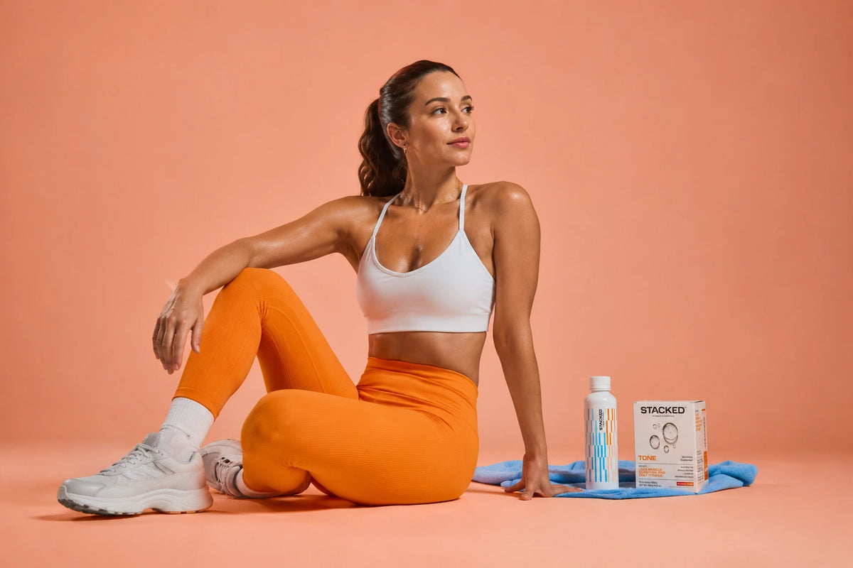 Fit woman in orange leggings and white sports bra sitting beside Stacked Tone, a creatine for women supplement, on a peach backdrop