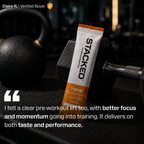 A Stacked TONE creatine and electrolyte stick pack standing upright on a gym floor between two black dumbbells, with a customer testimonial quote overlaid at the bottom